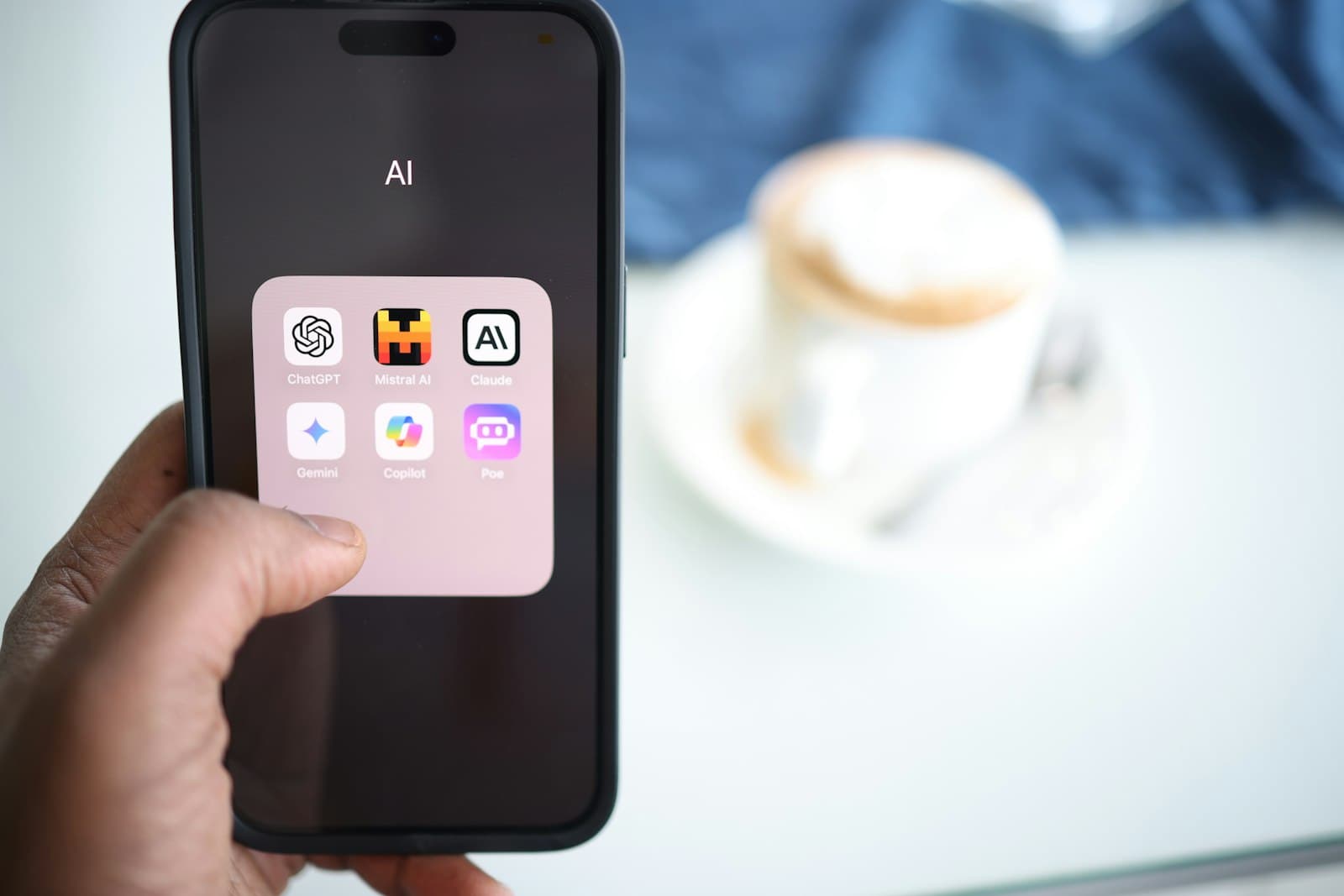 A hand holds a phone showing a folder of AI app icons including ChatGPT, Claude, Gemini, and Copilot